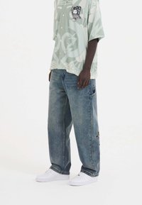 Short-sleeve, light green shirt with graphic print and buttons, paired with loose-fitting blue jeans featuring an embroidered patch and white sneakers.