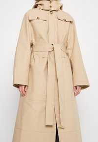 Trench-coat long beige avec manches larges, taille ceinturée, col boutonné et deux poches poitrine à rabat.