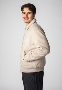 Beige Wildleder-Bomberjacke mit einem Reißverschluss auf der Vorderseite, seitlichen Taschen und gerippten Bündchen. Getragen über dunklen Jeans, mit einer lässigen Passform und weichem Stoff.