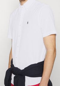 Vit kortärmad skjorta med knapp-up med texturerad tyg och en svart logotyp. En marinblå tröja är knuten runt midjan.