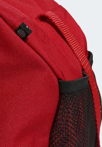 Mochila sintética roja con tela texturizada, que cuenta con un bolsillo lateral de malla negra y un lazo en la parte superior para llevarla. Los detalles clave incluyen líneas de costura.