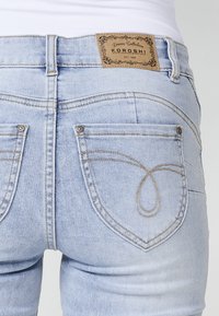 Light blue denim jeans with a dark stitched design. Features two rear pockets with decorative stitching and a tan leather tag at the waistband.