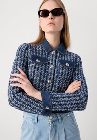 Veste en tweed bleu avec col et poignets en denim, ornée d'accents en boutons argentés et de deux poches sur la poitrine. Portée sur un jean en denim clair.