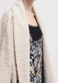 Beige, texturerad cardigan med sjalkrage, bärs över en svart tanktopp med ett silverpärlemönster. Mjuk och mysig look.