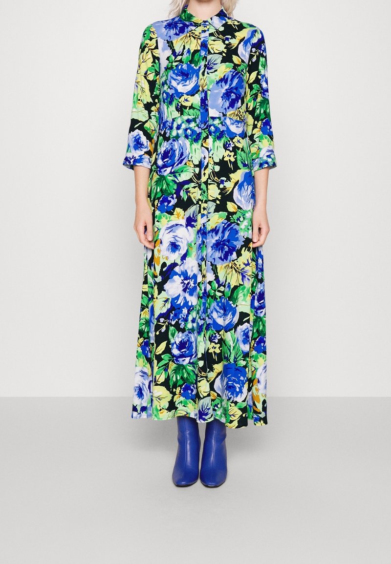 Blommig maxiklänning i blått, grönt och gult. Långa ärmar, knapplist fram och tyg med en glänsande yta. Kombineras med blå stövlar.