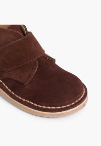 Zapato de ante marrón con una correa de velcro, punta redondeada y costuras contrastantes en tono claro a lo largo del borde de la suela.