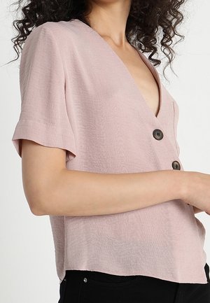 Kobieta ubrana w jasnoróżową bluzkę z krótkim rękawem i dekoltem w serek oraz dwa duże ciemne guziki, do tego czarne spodnie.