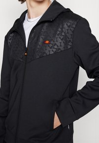 Black zip-up jacket with a textured black upper panel, side pockets, and an embroidered logo. Smooth fabric with a modern fit.