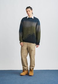 Πλεκτό πουλόβερ με μπλε, πράσινες και γκρι ρίγες, φορεμένο πάνω από ένα λευκό πουκάμισο. Παντελόνια cargo σε μπεζ και τακούνια σε μαύρο χρώμα ολοκληρώνουν την εμφάνιση.