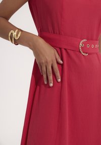 Czerwona sukienka z szerokim paskiem i metalową okrągłą klamrą, w zestawieniu ze złotą bransoletą o falistym wzorze, prezentowana na neutralnym tle.