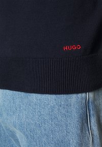 Námořnický pletený svetr s žebrovanými manžetami a lemováním. Obsahuje malé červené logo "HUGO" poblíž spodní části. Vybavený s lehce modrými džíny.