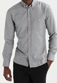 Camisa gris de botones con mangas largas, un único bolsillo en el pecho, botones en contraste y un acento de cuero de marca en el dobladillo.