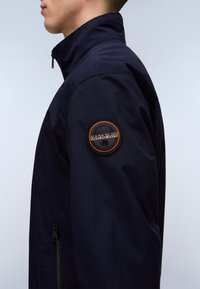 Boční pohled na osobu nosící námořnicky modrou bundu s kapsou na zip a kulatým černo-oranžovým logem Napapijri na horní části paže.