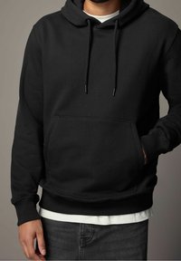 Homme portant un sweat à capuche noir sur une chemise blanche, une main dans la poche avant et un jean sombre visible en bas.