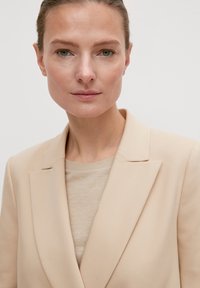 Beige Blazer mit einem strukturierten Reversdesign, kombiniert mit einem hellbeigen Pullover darunter. Glattes Material mit einem minimalistischen, klaren Look.