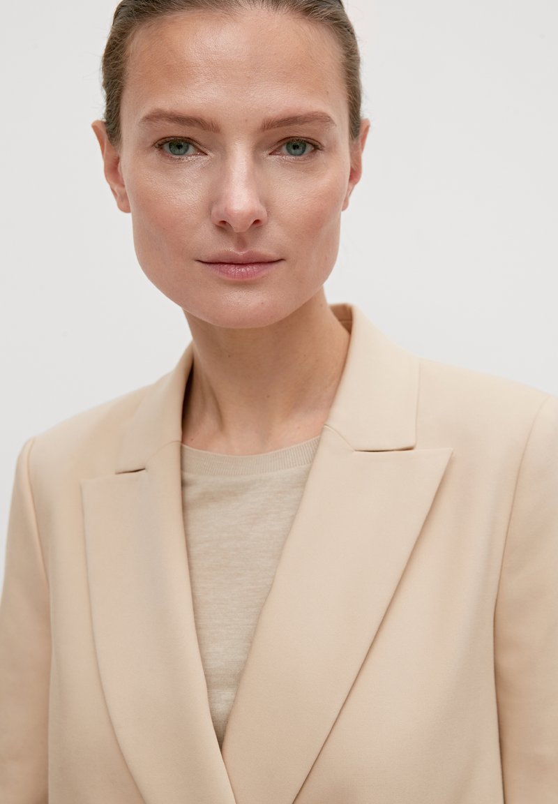 Blazer beige avec un design de revers structuré, associé à un pull beige clair en dessous. Tissu doux avec une apparence minimaliste et épurée.