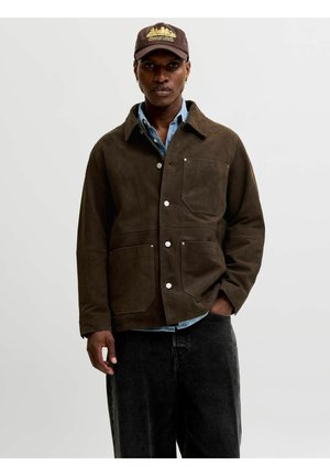 Homme portant une veste en daim marron, une chemise en denim bleu, un jean noir et une casquette marron avec une broderie dorée de la silhouette de la ville.