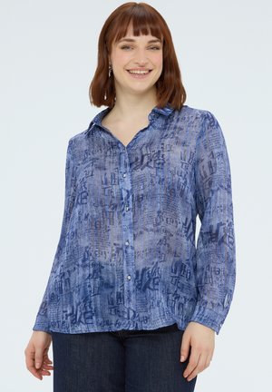 Lächelnde Frau mit schulterlangem kastanienbraunem Haar, die eine blaue, gemusterte Bluse mit Knöpfen und eine dunkle Jeans trägt, vor einem einfarbigen Hintergrund.