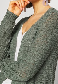 Žena v zeleném otevřeném pleteném svetru přes bílý top, dotýkající se brady, s levandulovými kruhovými náušnicemi a krátkými hnědými vlasy.