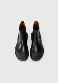 Une paire de bottines Chelsea en cuir noir avec des orteils arrondis, des panneaux latéraux élastiques, des languettes de tirage et des semelles crantées sur un fond blanc uni.