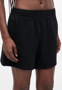 Personne portant un short noir à taille élastique avec poches latérales, montrée de l'abdomen à la mi-cuisse sur un fond uni.