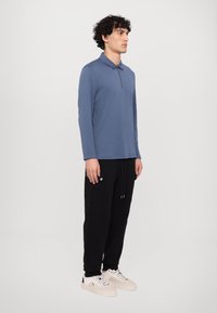 Langærmet blå polo med en enkelt lynlås ved kraven, parret med sorte joggingbukser med sidelommer og snoretræk i livet.
