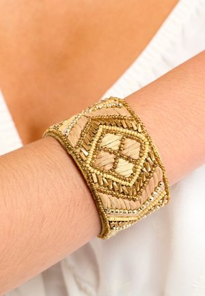 Bracelet manchette tissé avec motifs géométriques dans des tons beiges et dorés. Comprend des perles métalliques et des fils texturés, mettant en valeur des détails de design complexes.