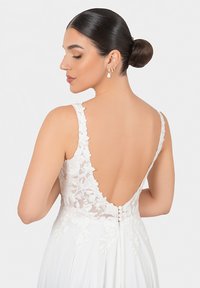 Abito da sposa elegante con un profondo design in pizzo sulla schiena, dettagli in pizzo floreale e una gonna bianca fluente con bottoni decorativi.