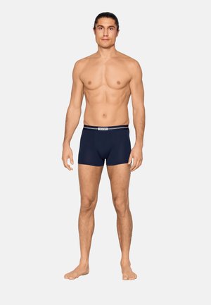 Uomo senza maglietta con capelli scuri, indossa boxer blu navy, in piedi a piedi nudi contro uno sfondo chiaro e semplice.