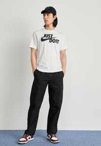 Hvid Nike t-shirt med "Just Do It" trykt i sort, sorte cargo bukser og røde og hvide sneakers. Modellen bærer en sort kasket.