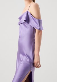 Kobieta w jedwabnej lawendowej sukience bez ramion z falbanką przy dekolcie i rozporkiem po boku, pokazana od ramion do połowy uda.