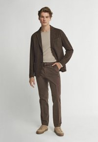 Brauner strukturierter Blazer über einem beigen Hemd, kombiniert mit braunen Hosen und beigen Sneakern. Der Blazer hat eine einzelne Tasche und einen Knopfverschluss.