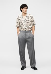 Νεαρός άνδρας στέκεται με τα χέρια στις τσέπες, φοράει μπεζ κοντομάνικο πουκάμισο με σχέδια, γκρι παντελόνι και μαύρα παπούτσια σε λευκό φόντο.