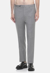 Uomo che indossa pantaloni formali grigio chiaro e mocassini in pelle nera, in piedi davanti a uno sfondo bianco semplice.