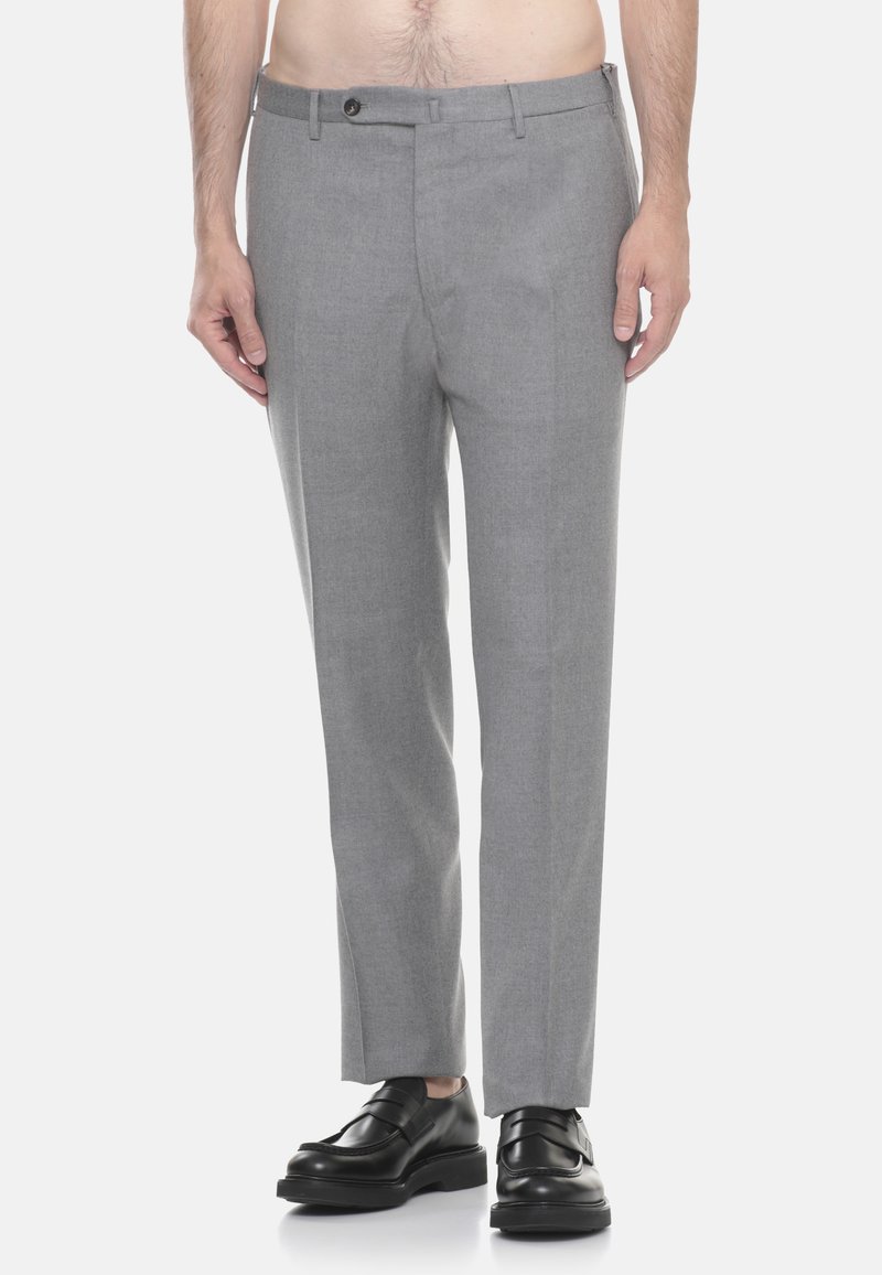 Uomo che indossa pantaloni formali grigio chiaro e mocassini in pelle nera, in piedi davanti a uno sfondo bianco semplice.