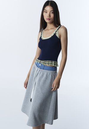 Mujer joven con cabello largo y oscuro, vistiendo una camiseta de tirantes azul marino acanalada, una camiseta interior a rayas en capas, cinturilla azul y falda gris con cordón ajustable.