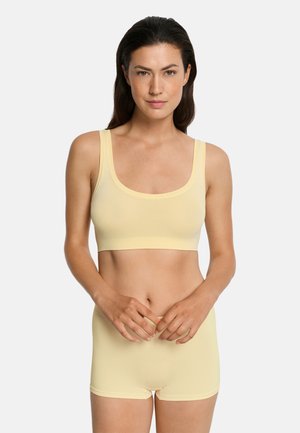 Kobieta o ciemnych włosach, ubrana w jasnopomarańczowy bezszwowy krótki top i pasujące do niego szorty, stojąca na tle jednolitego jasnego tła, z rękami złączonymi.