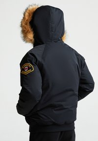 Giacca nera imbottita con cappuccio foderato in pelliccia, caratterizzata da una toppa gialla e rossa sulla spalla sinistra. Texture liscia, lunghezza corta.