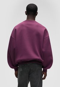 Felpa oversized viola con spalle scese, polsini a coste e una texture liscia. Abbinata a jeans grigi scuri con un logo sottile in vita.