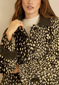 Fekete és krémszínű foltos szőrme kabát texturált felülettel, sötét gallérral, nagy gombokkal és övvel díszített ujjal.