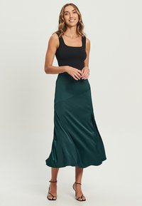 Satin-türkiser Rock mit fließendem Design, der einen diagonalen Naht und seitliche Schlitze aufweist, kombiniert mit einem engen schwarzen Top und Riemchensandalen.