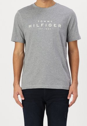 Mężczyzna ubrany w szarą koszulkę z krótkim rękawem i okrągłym dekoltem z napisem "Tommy Hilfiger EST. 1985" na piersi oraz ciemne spodnie.