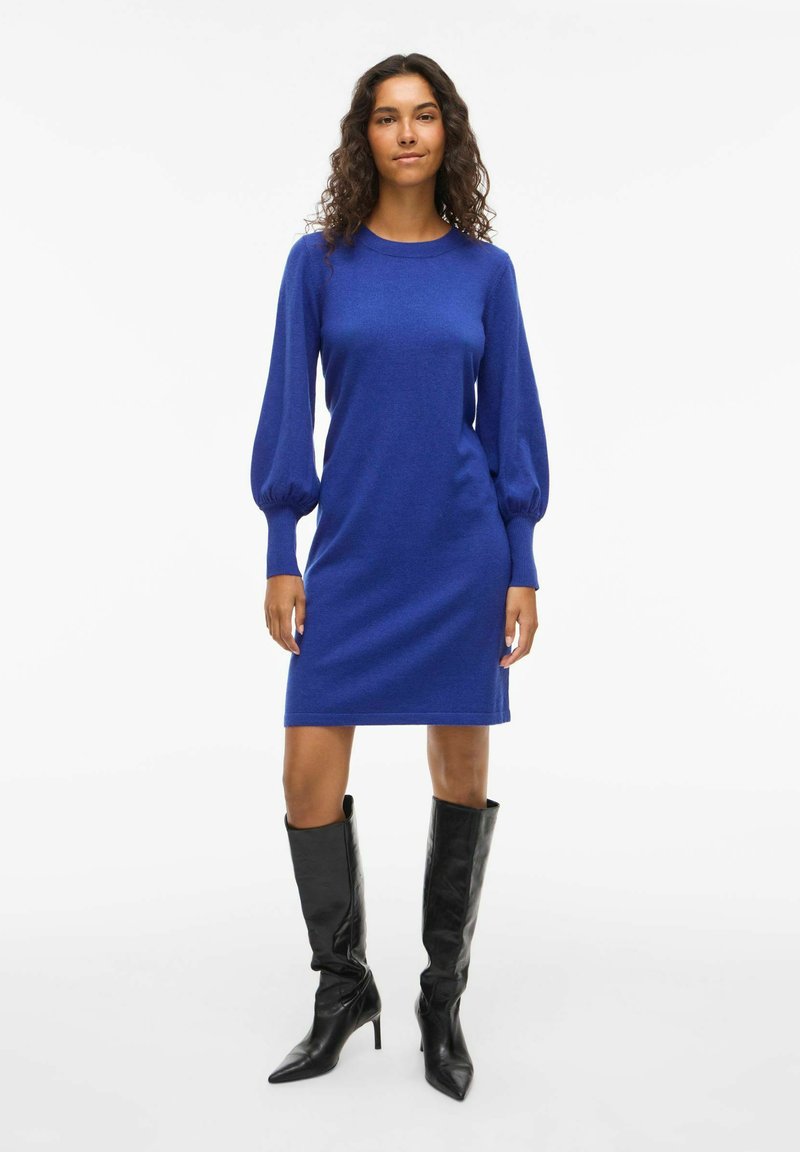 Vestito lungo in maglia a maniche lunghe blu con corpino aderente e maniche a palloncino, abbinato a stivali neri in pelle con tacco alto. Texture liscia e design classico.