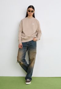 Felpa beige con scollo a girocollo e piccolo logo, abbinata a jeans baggy blu sbiaditi con orlo strappato e sneakers a scacchi.