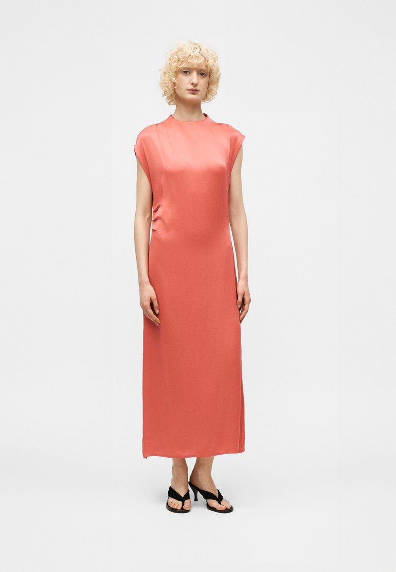 Ξανθός άνθρωπος στέκεται, φορώντας ένα μακρύ χωρίς μανίκια κοραλί φόρεμα και μαύρα σανδάλια με τακούνι μπροστά σε ένα απλό φωτεινό φόντο.