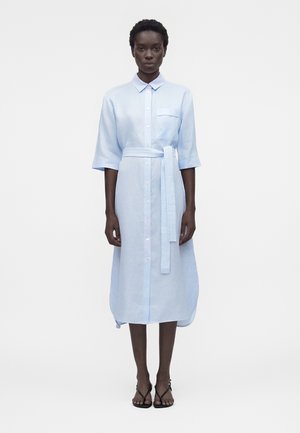 Femme debout portant une robe chemise midi bleu clair avec col, boutons devant, poche poitrine, ceinture nouée à la taille, et sandales noires.