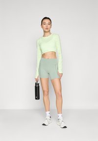 Jasnozielony, krótką rękawą top z subtelnym logo, połączony z jasnozielonymi szortami. Model trzyma czarną butelkę na wodę, nosząc białe sneakersy.