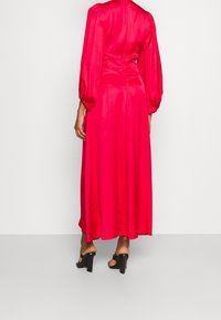 Kvinna klädd i en långärmad, klarrosa satinklänning med puffärmar och svarta högklackade sandaler, stående mot en naken vit vägg.