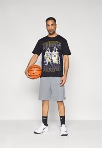 Μαύρο t-shirt με γραφική εκτύπωση που απεικονίζει καλαθοσφαιριστές, συνδυασμένο με γκρι σορτς, λευκά αθλητικά παπούτσια και κρατώντας πορτοκαλί μπάλα μπάσκετ.