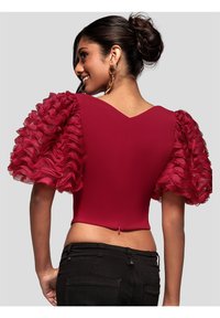 Rød tætsiddende top med puf-agtige, gennemsigtige flæseærmer, dyb V-udskæring bagpå og lynlåsbaglukning. Bærtes med sorte bukser.